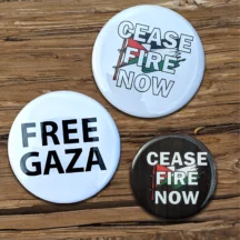 wood background with three buttons laying atop. the highest button says "cease fire now" with the Palestinian flag behind it. button left of center says "free Gaza" in all black caps with a white background. The smaller button on the right bottom says "cease fire now" on a black background with the Palestinian flag behind.